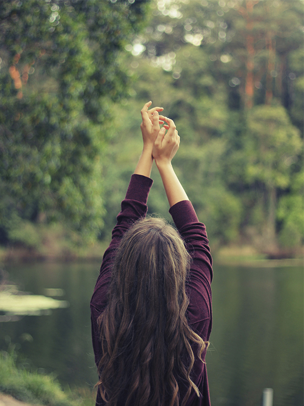 Rückansicht einer Frau die an einem See steht und die Arme über den Kopf streckt.Foto: Christopher Campbell via Unsplash