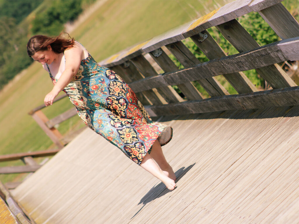 Tess tanzt im Sonnenschein in einem bunten Kleid über eine Holzbrücke.