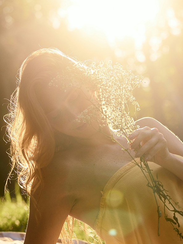Gegenlichtportrait einer jungen Frau in einem gelben Kleid, die mit weißen Blumen einen Teil ihres Gesichts verdeckt.